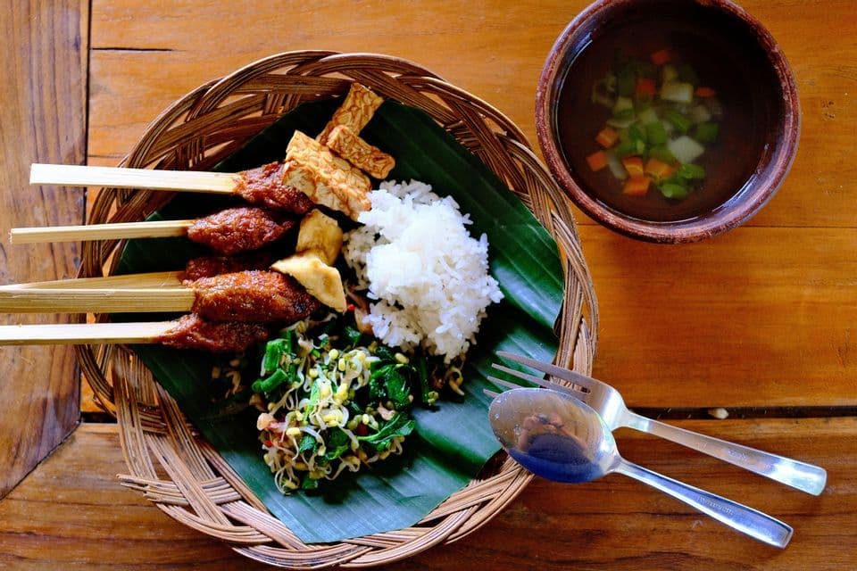 Eine Platte mit Satay-Spießen, Tempeh, Reis und Salat auf einem geflochtenen Teller, dazu Gemüsesuppe.