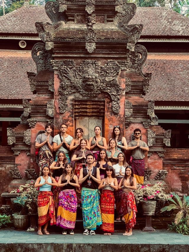 Un groupe WeRoad en voyage, vêtu de sarongs colorés, pose les mains jointes en signe de prière devant un temple de pierre orné.