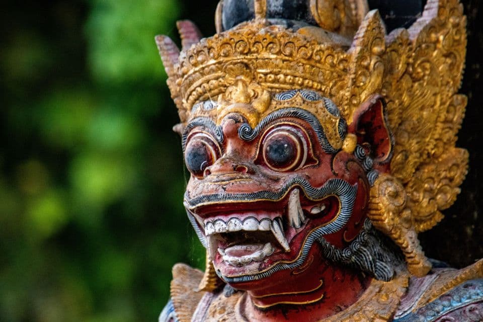 Nahaufnahme einer traditionellen asiatischen Wächterstatue mit rotem Gesicht, entblößten Reißzähnen und einer aufwendigen goldenen Kopfbedeckung.