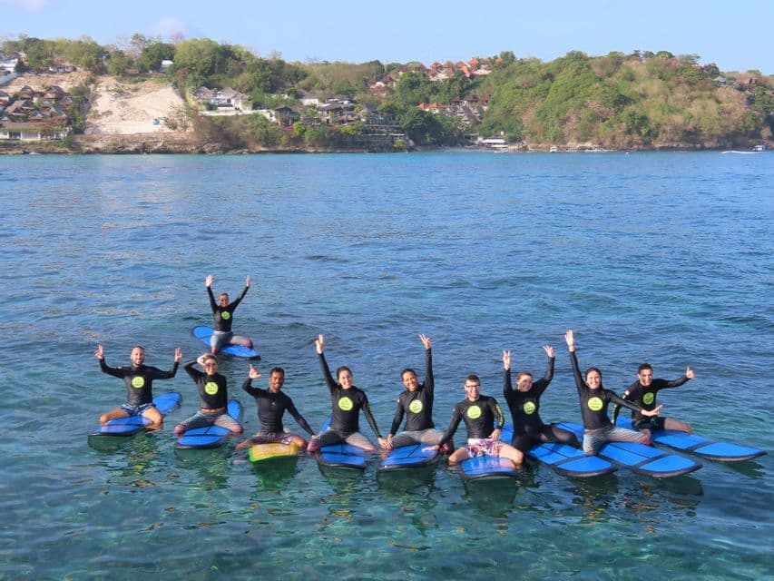 A WeRoad group trip in wetsuits posing on blue surfboards in the clear sea, with a green coastline in the background.