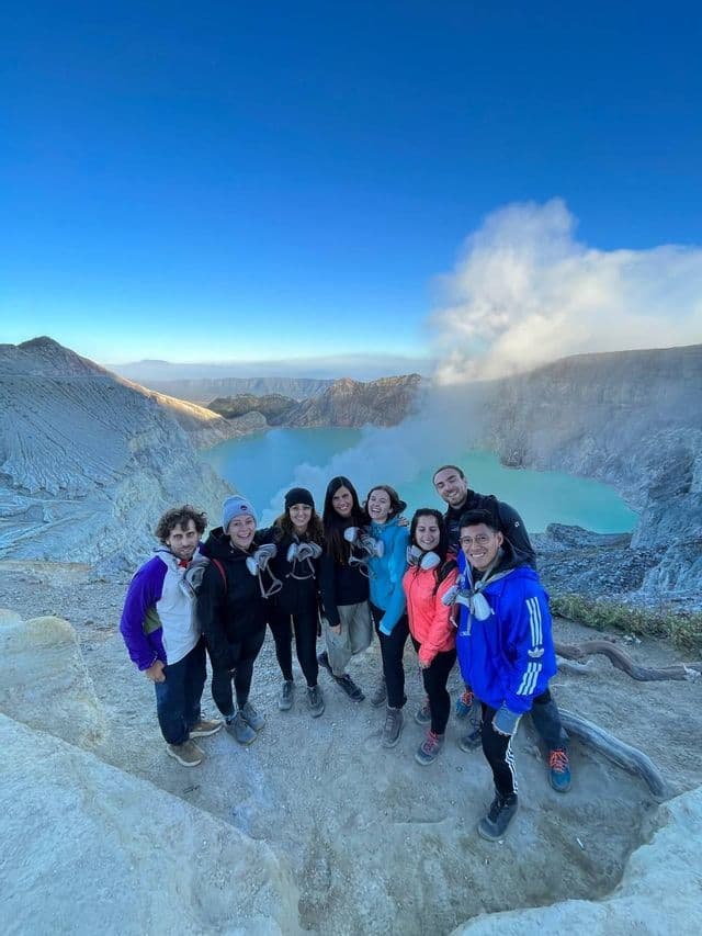 A WeRoad group trip poses for a photo on the edge of a volcanic crater, with a bright turquoise lake in the background.