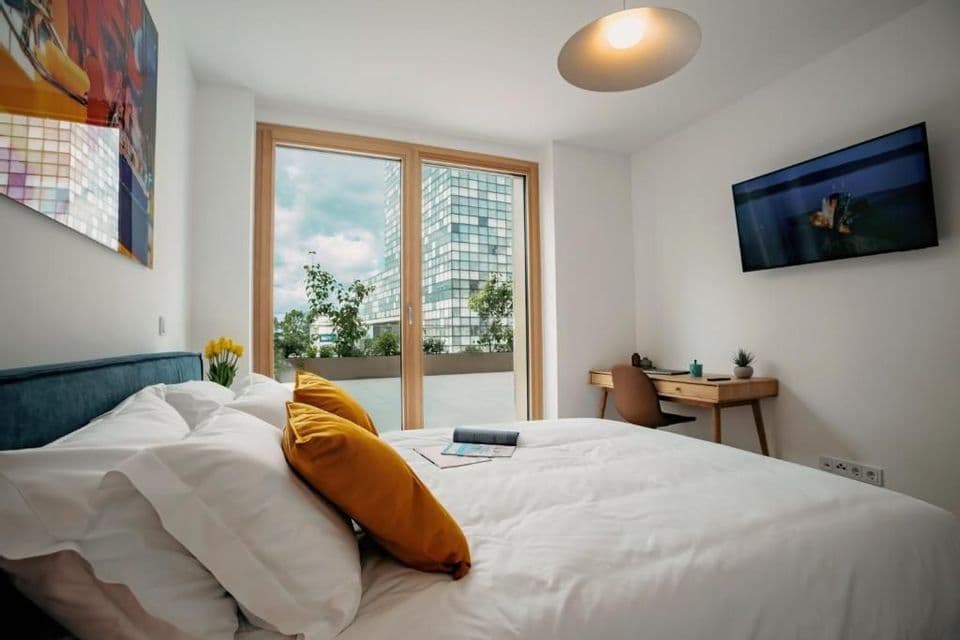 Interno di una camera da letto moderna con un letto bianco, una scrivania e una porta a vetri che si apre su un balcone con vista città.