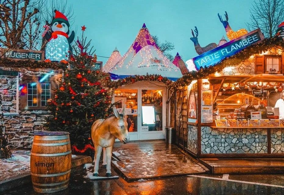 Un mercatino di Natale al tramonto, con decorazioni illuminate, tra cui una statua di pinguino e una di asino, accanto a bancarelle festive su una strada bagnata.