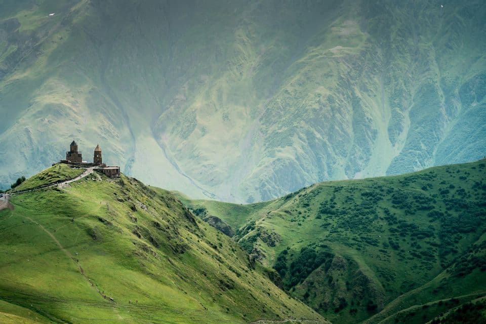 Un'antica chiesa in pietra con due torri si erge in cima a una rigogliosa collina verde, con uno sfondo di vaste montagne a strati sotto un cielo velato.