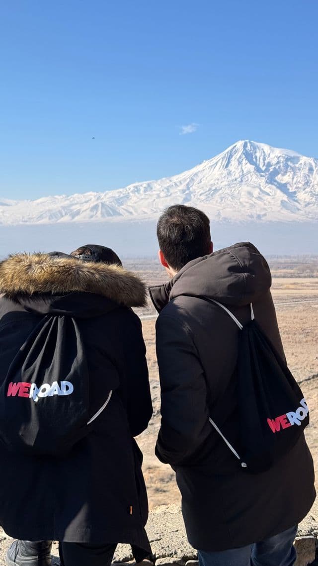 Due persone di un viaggio di gruppo WeRoad, viste da dietro, guardano una montagna innevata in lontananza sotto un cielo sereno.