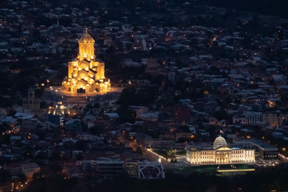 Una cattedrale illuminata e un grande edificio con una cupola si affacciano su un paesaggio urbano notturno.