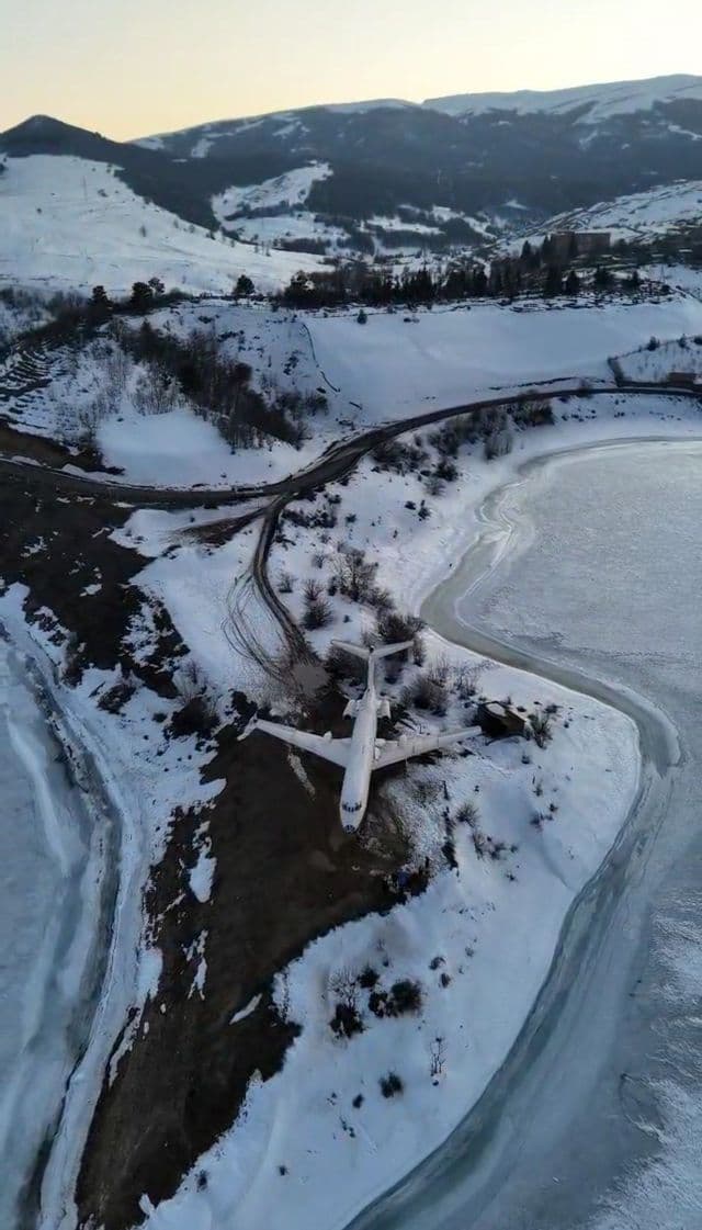 Veduta aerea di un aereo bianco sulla sponda innevata di un lago ghiacciato, circondato da colline innevate.