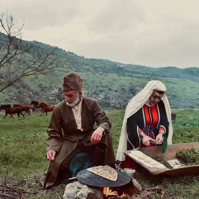 Un uomo e una donna in abiti tradizionali cucinano pane piatto su un falò in un campo erboso con cavalli sullo sfondo.