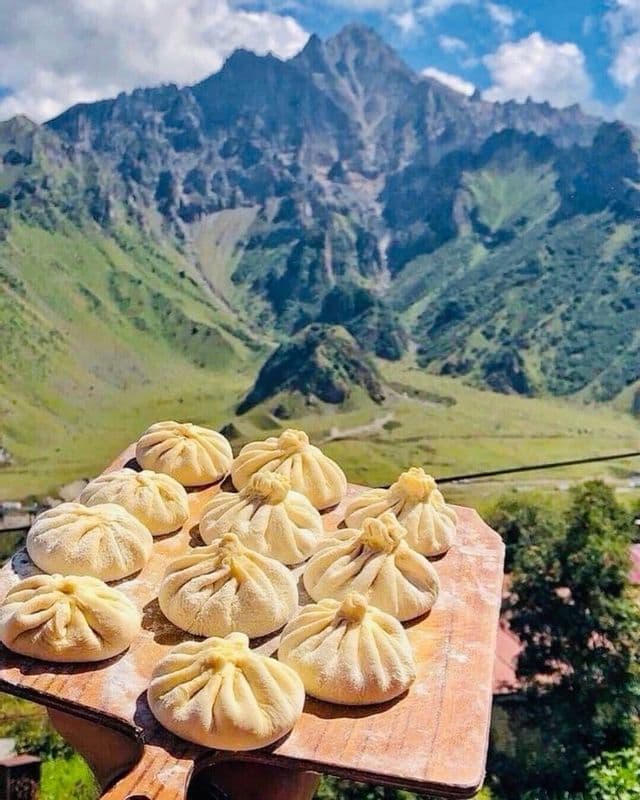 Una tavola di legno con gnocchi crudi è presentata davanti a una grande catena montuosa verde sotto un cielo parzialmente nuvoloso.