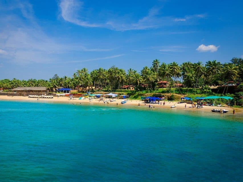 Un'ampia vista di una spiaggia tropicale con acqua turchese, una riva sabbiosa con persone e una fila di palme sotto un cielo azzurro.
