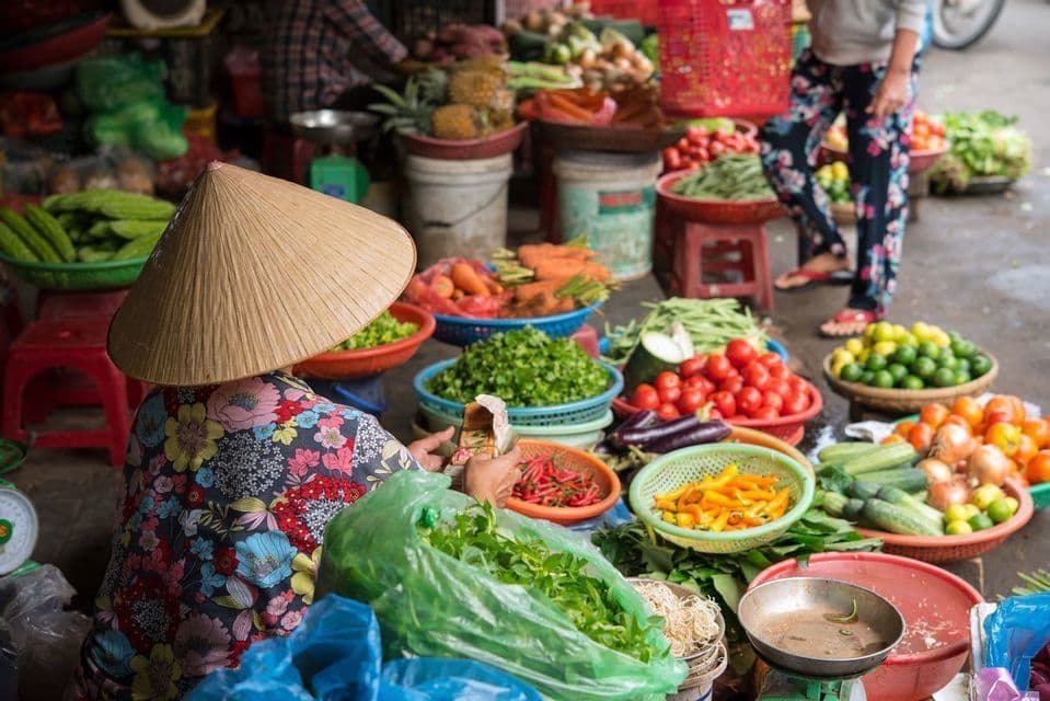 Vu de dos, un vendeur portant un chapeau conique est assis entouré de paniers de légumes frais sur un étal de marché en plein air.