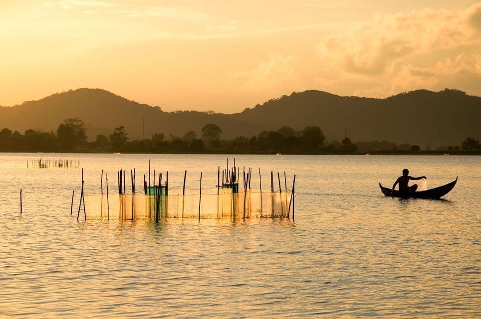 A silhouette of a fisherman casts a net from a boat on a calm lake with mountains in the background during a golden sunset.