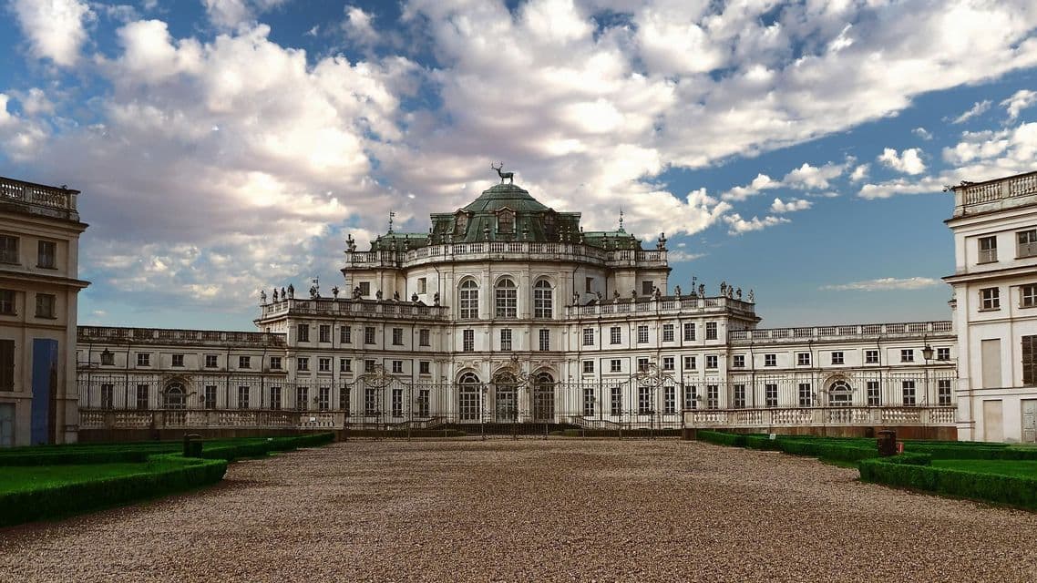 Un gran palacio barroco blanco con una estatua de ciervo en su cúpula central se alza detrás de un patio de grava bajo un cielo nublado.