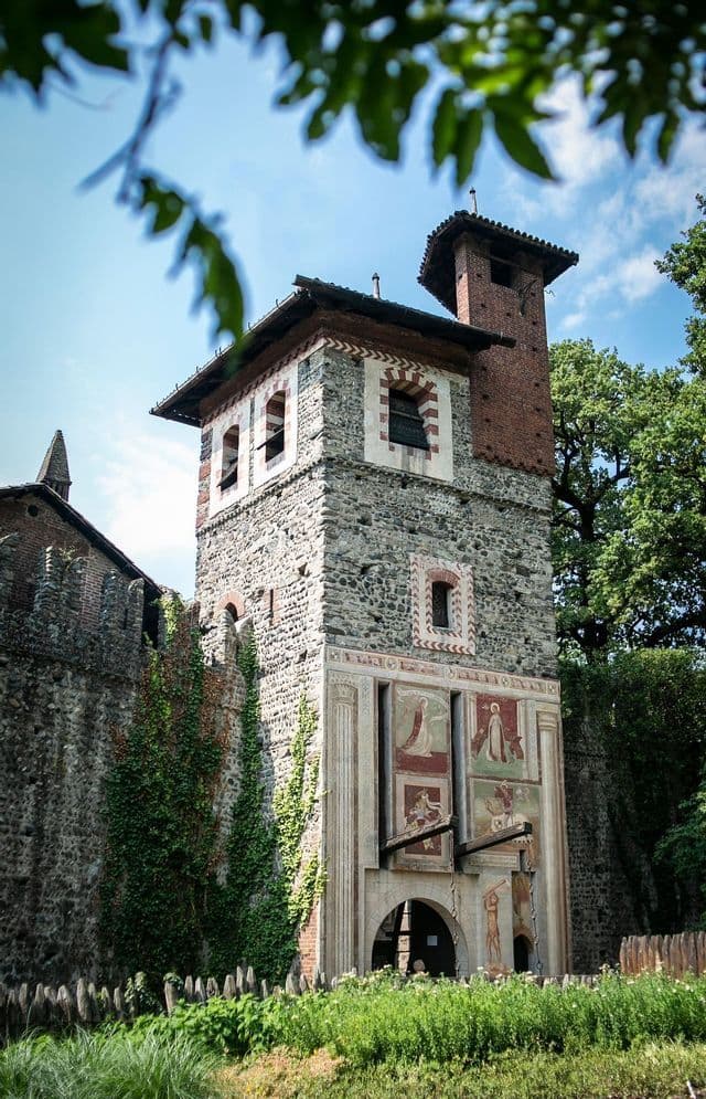 Una torre de piedra y ladrillo de un castillo medieval, decorada con frescos, vista entre hojas de árboles contra un cielo azul.