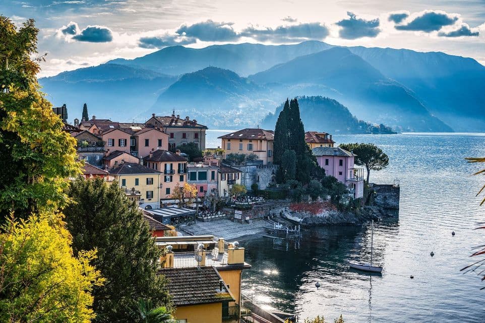 Una vista aérea de un colorido pueblo a orillas del lago anidado al pie de grandes montañas brumosas bajo un cielo parcialmente nublado.