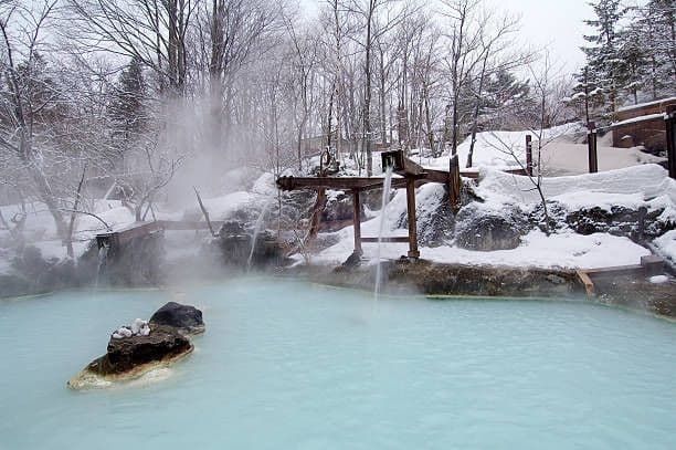 A wooden aqueduct pours water into a steaming, milky-blue hot spring in a snowy, wooded landscape.
