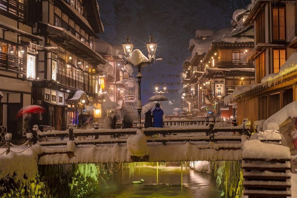 Ein verschneites Dorf bei Nacht, mit traditionellen Gebäuden, einer Brücke über einen Kanal und beleuchteten Laternen.
