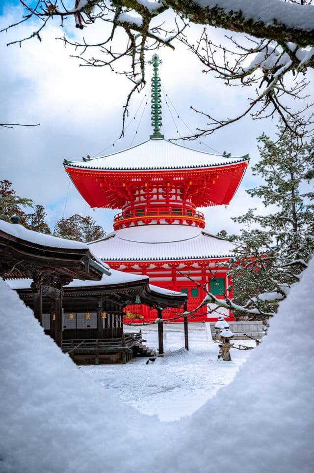 A red Japanese pagoda with snow-covered roofs and grounds, viewed through a natural frame of snow-laden tree branches.