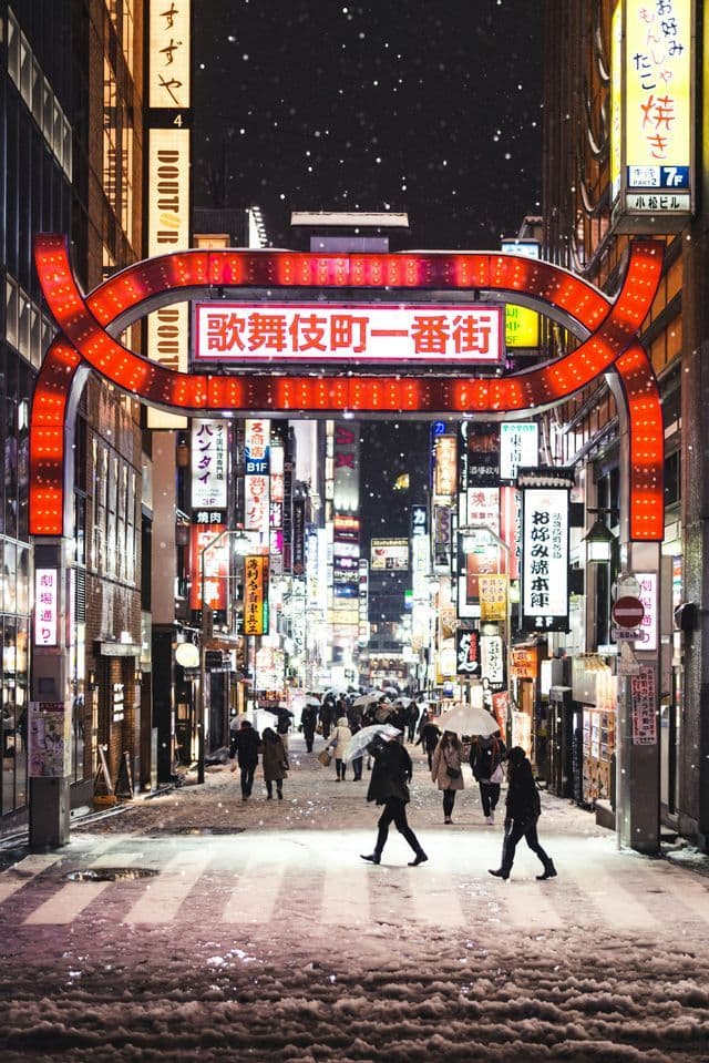 People walk on a snowy city street at night under a large red archway and glowing signs with Asian characters.