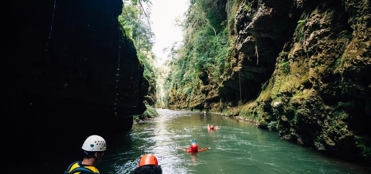 Un gruppo WeRoad in viaggio discende un fiume attraverso uno stretto e verdeggiante canyon, indossando caschi e giubbotti di salvataggio.