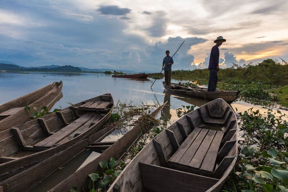 Deux hommes se tiennent sur un bateau en bois sur un lac calme, entourés d'autres bateaux et de plantes aquatiques, pendant un coucher de soleil nuageux.