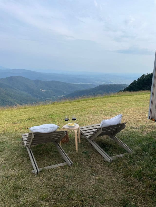 Due sedie a sdraio in legno e un tavolino con bicchieri da vino si trovano su un pendio erboso che si affaccia su una catena montuosa.