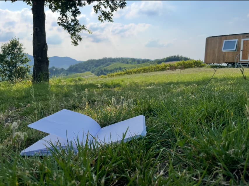 Un quaderno aperto a righe giace in un campo erboso con una casetta e verdi colline ondulate sullo sfondo.