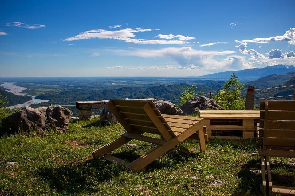 Sedie a sdraio in legno su un punto panoramico erboso con vista su un'ampia valle fluviale e montagne sotto un cielo parzialmente nuvoloso.