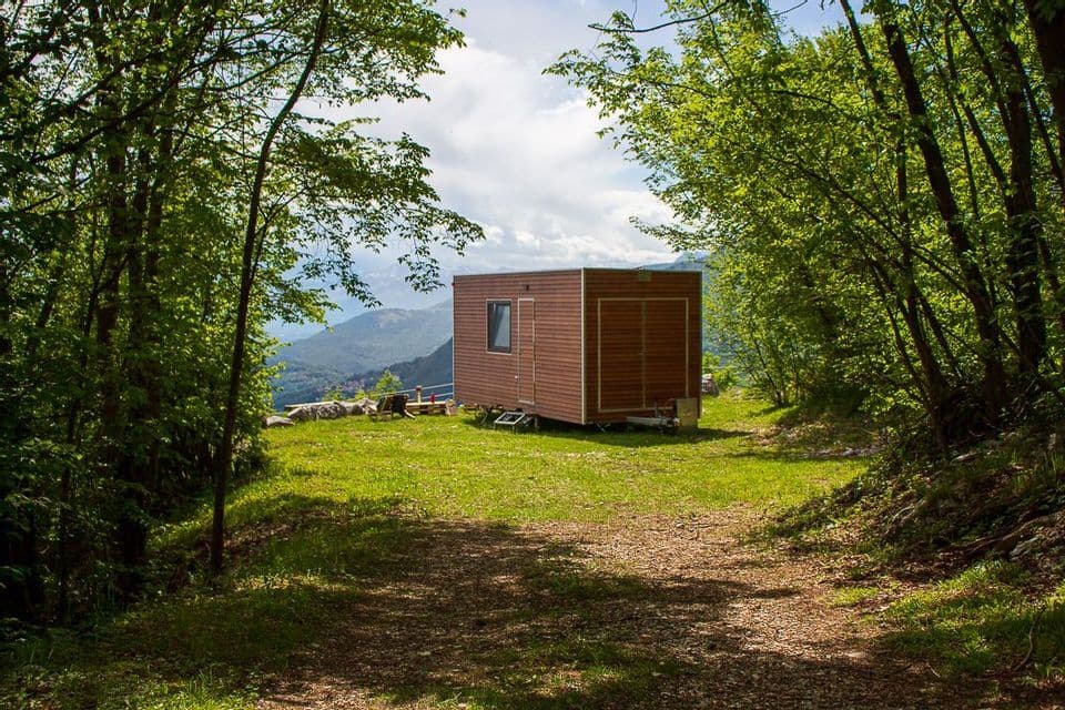 Una piccola casa di legno si trova in una radura erbosa in una foresta, con vista su montagne distanti.