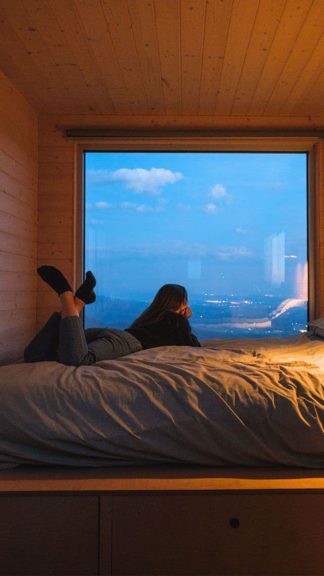 Una donna giace su un letto in una stanza di legno, guardando attraverso una grande finestra una città al crepuscolo.