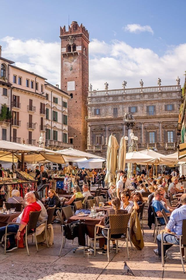 Una multitud de personas se sienta en cafés al aire libre y explora los puestos del mercado en una soleada y histórica plaza europea con una prominente torre de ladrillo.