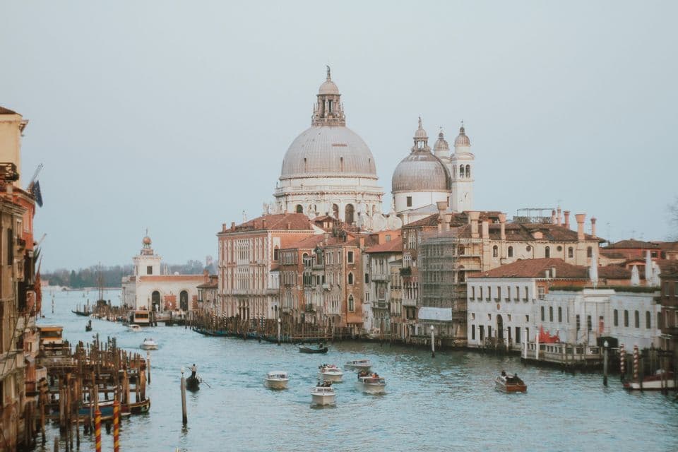 Barcos y góndolas navegan por un amplio canal bordeado de edificios históricos y una gran basílica con cúpula bajo un cielo encapotado.