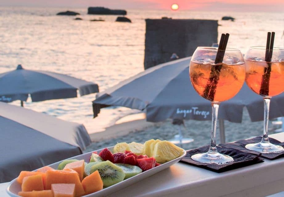 Due cocktail all'arancia e un piatto di frutta fresca su un davanzale che si affaccia su una spiaggia con ombrelloni al tramonto.