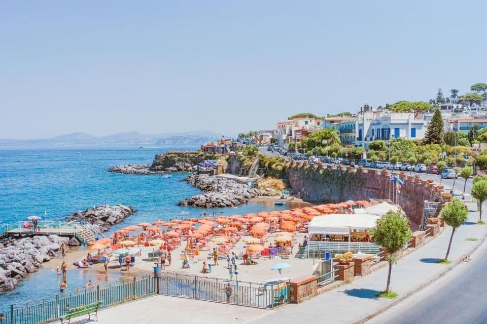 Una spiaggia affollata con ombrelloni arancioni è incastonata in una caletta rocciosa accanto a una località costiera in una giornata di sole.