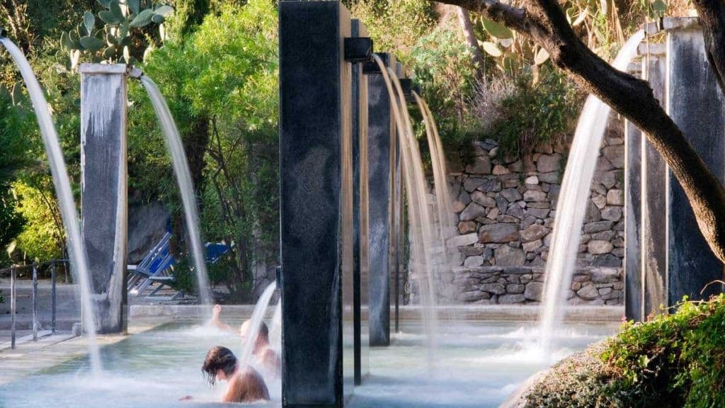 Persone che si rilassano in una piscina termale all'aperto con acqua che scende da grandi fontane di pietra circondate da una vegetazione lussureggiante.