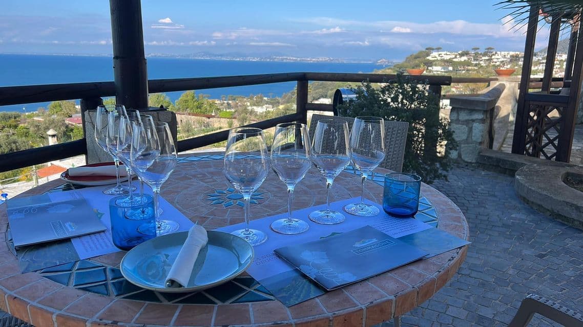 Un tavolo a mosaico è apparecchiato con bicchieri da vino vuoti per una degustazione, affacciato su un vasto mare blu e una costa distante.