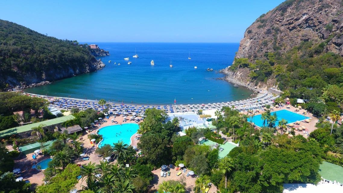 Vista aerea di un resort balneare con piscine accanto a una spiaggia a mezzaluna affollata in una cala.