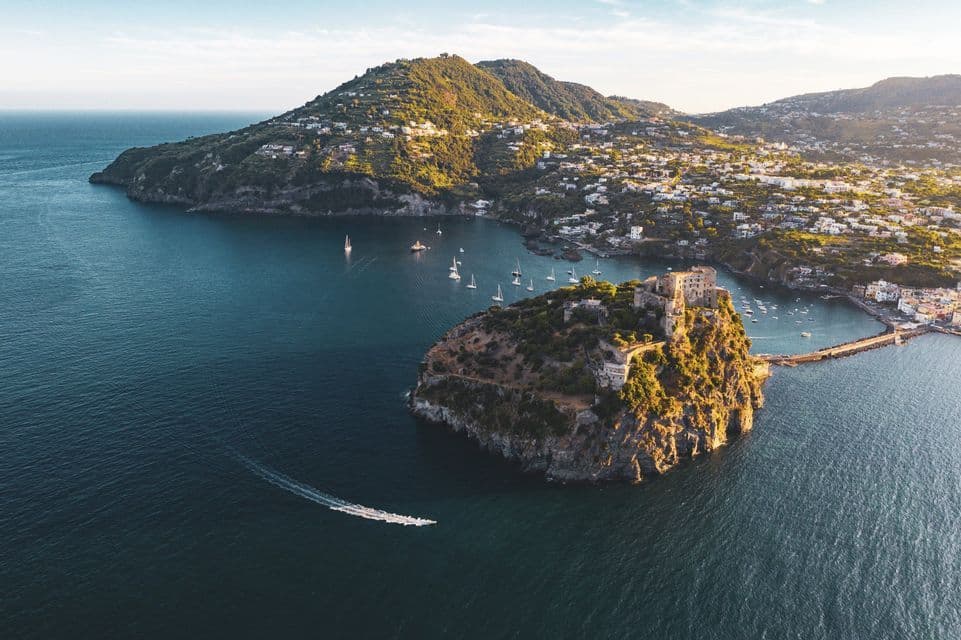 Veduta aerea di un castello su un isolotto roccioso collegato da un ponte a una città sulla terraferma, con barche a vela nella baia.