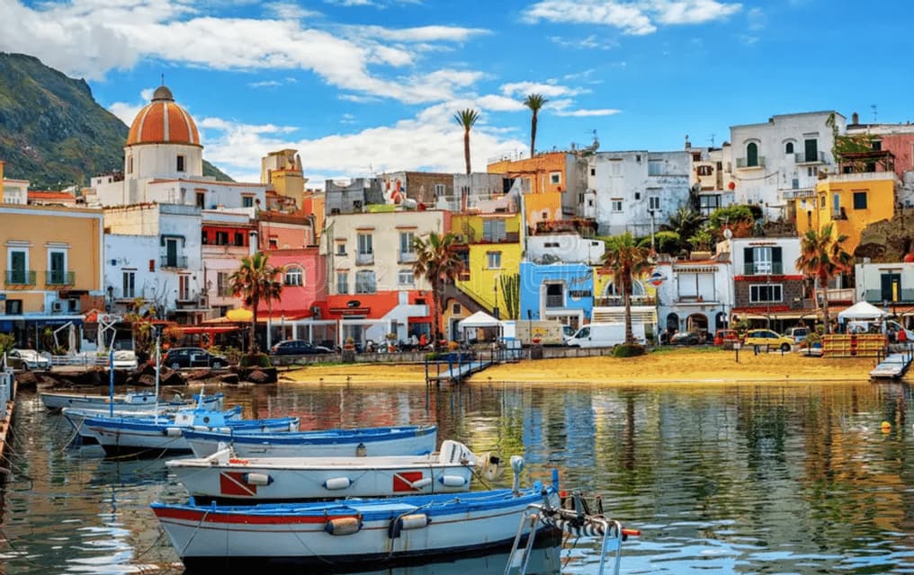 Barche sono ormeggiate in un porto di fronte a una colorata cittadina di mare con una chiesa a cupola e una spiaggia sotto un cielo blu.