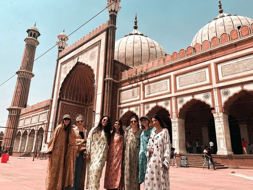 Un voyage de groupe WeRoad de sept femmes posant dans une cour devant une grande mosquée en grès rouge avec des dômes blancs.