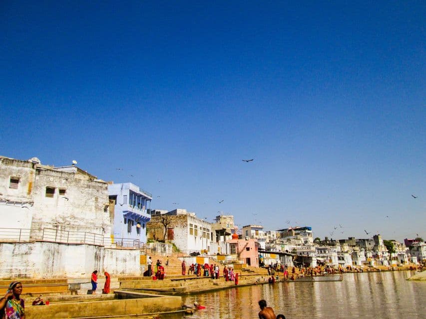 Des personnes aux tenues colorées se rassemblent sur des ghats en pierre le long d'un front de mer, avec un paysage urbain dense sous un ciel bleu éclatant.
