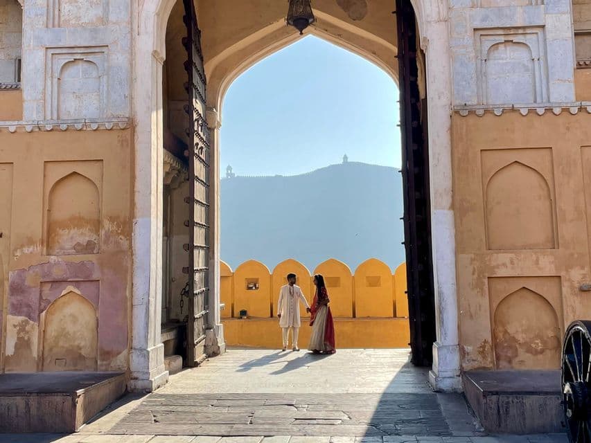 Una pareja con atuendos tradicionales indios se encuentra dentro de un gran portal arqueado, con una colina lejana visible a través de la abertura.