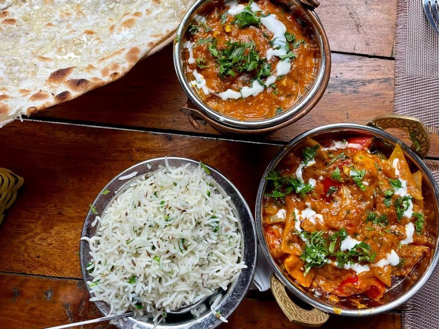 Draufsicht auf zwei Schüsseln indisches Curry, eine Schüssel gewürzten Reis und ein Stück Naan-Brot auf einem Holztisch.