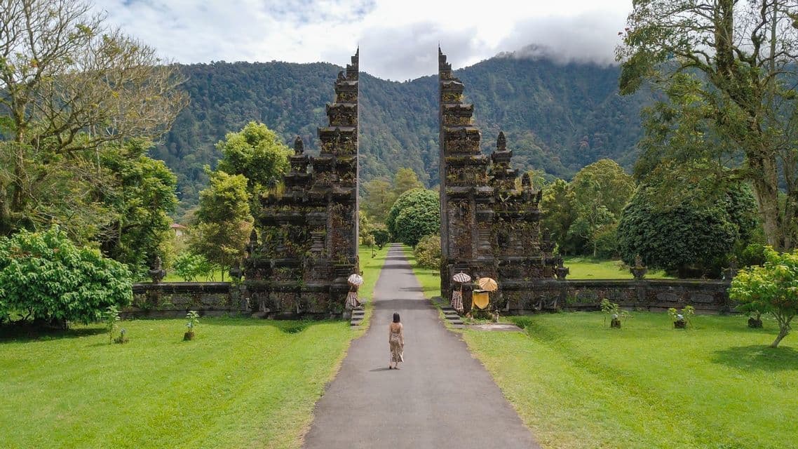 A person walks on a path leading through a tall, ornate stone gate, with lush trees and forested mountains in the background.