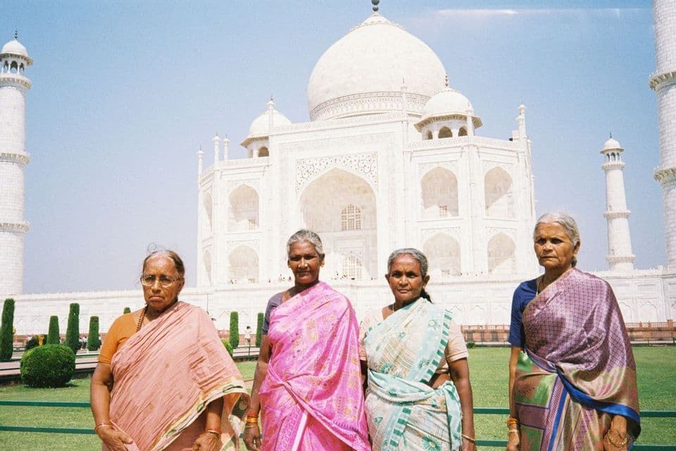 Cuatro mujeres mayores con coloridos saris de pie en un césped verde frente a un gran mausoleo de mármol blanco y cúpula central.