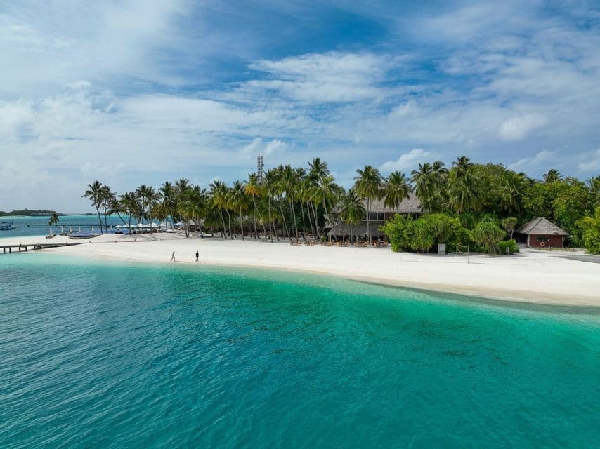 Una vista aérea de una isla tropical con playa de arena blanca, agua turquesa y palmeras bajo un cielo parcialmente nublado.
