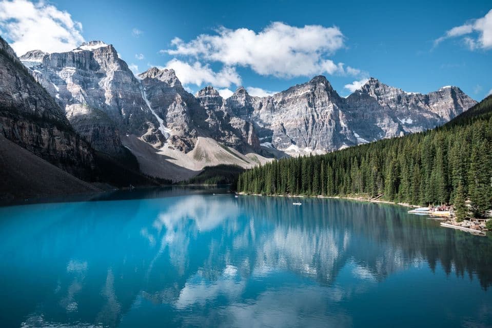 Ein leuchtend blauer Alpensee spiegelt schneebedeckte Felsenberge und einen dichten Kiefernwald unter einem blauen Himmel mit vereinzelten Wolken wider.