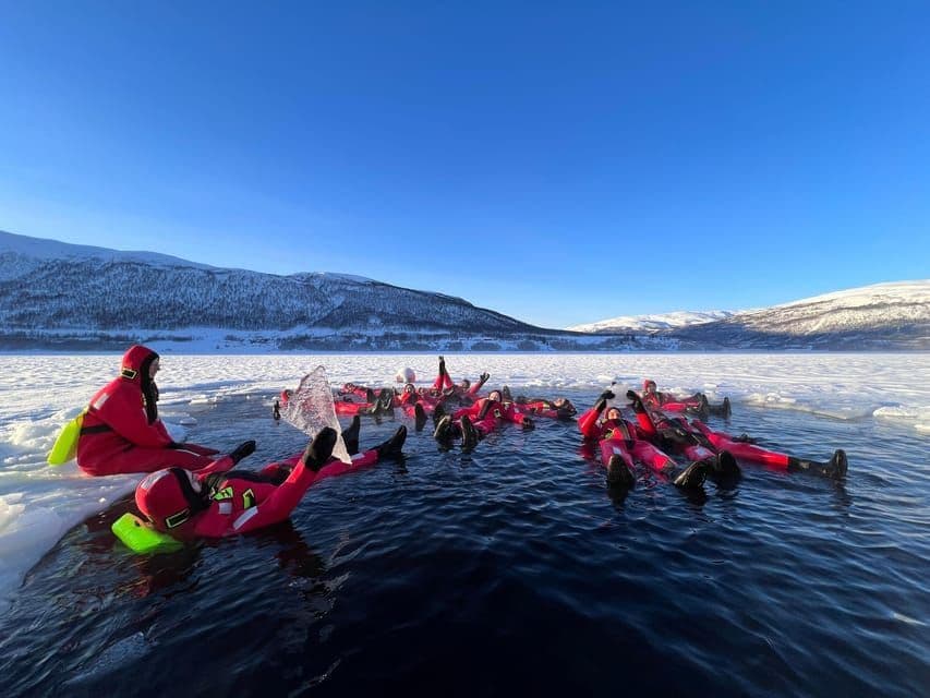 A WeRoad group trip wearing red thermal suits floats in the icy water of a frozen lake, surrounded by snowy mountains.