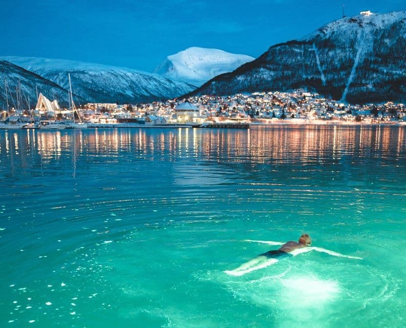 A person floats on their back in glowing turquoise water at night, with a snowy, illuminated town and mountains in the background.