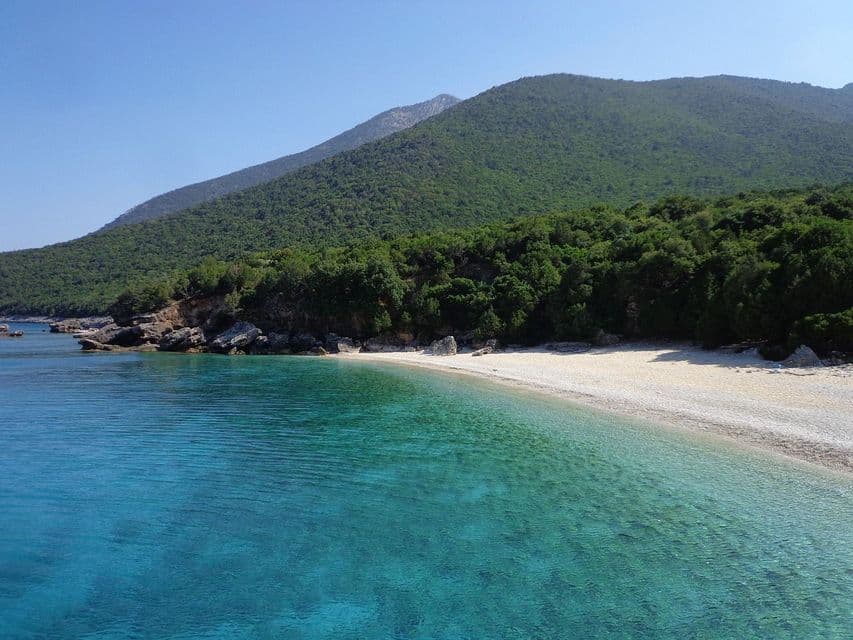 Acque cristalline e turchesi lambiscono una spiaggia isolata di ciottoli bianchi ai piedi di una montagna lussureggiante e alberata, sotto un cielo azzurro.